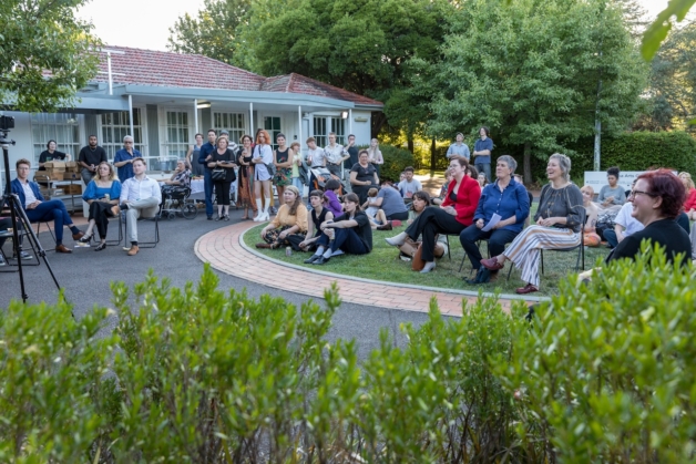 Photo of people sitting on chairs and grass in the Manuka Arts Centre gardens listening to the opening speech on a summers early evening