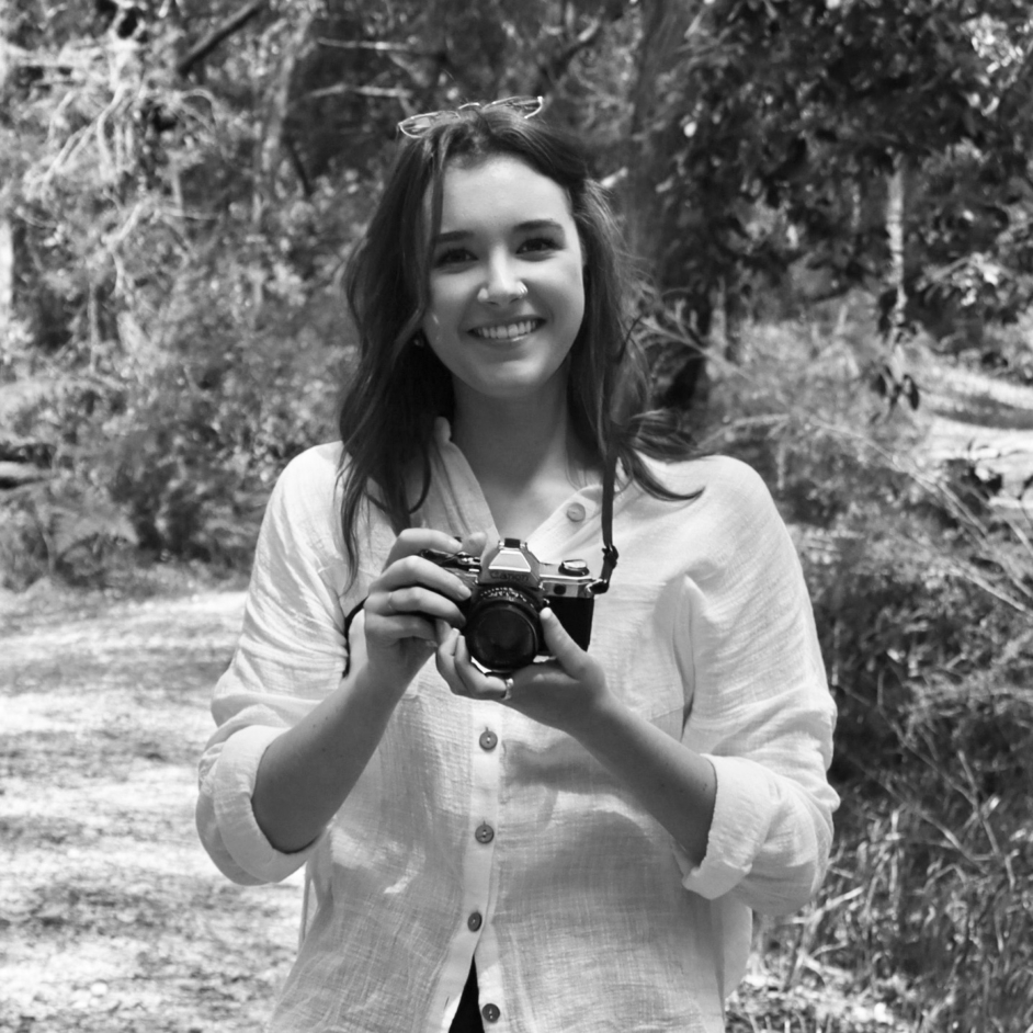 Black and white photograph of a young woman smiling and holding a film camera in front of her chest against a bush backdrop