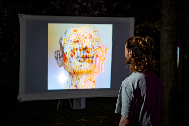 Photograph behind a man looking at a projection screen showing a warped digital rendering of a face in yellow, red and blue.