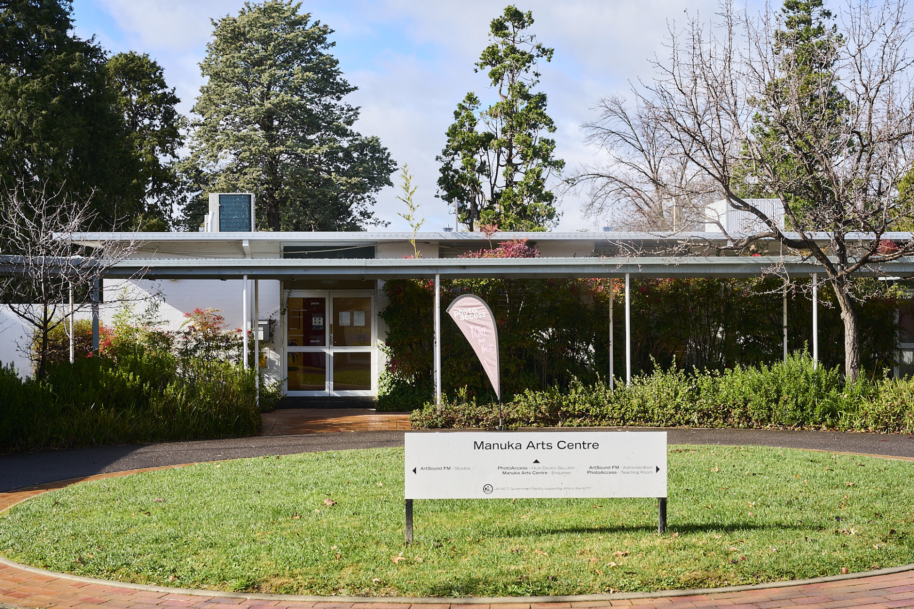 Photograph at the front of the white single story photo access building with a grassy roundabout and a sign saying "Manuka Arts Centre" in front on a blue sunny day.