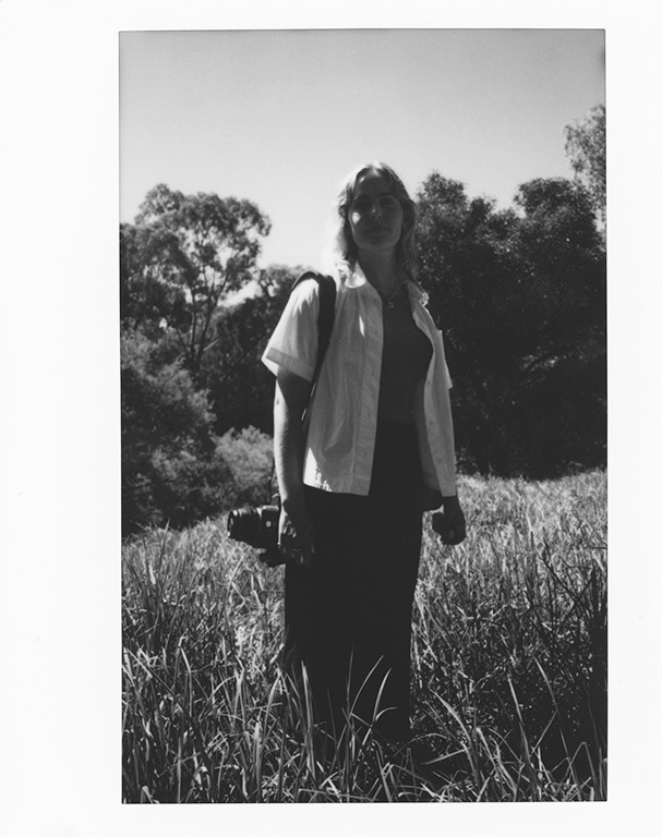 Black and white photograph of a woman with a camera in her hand standing in a field of long grass, with open sky and trees in the background.