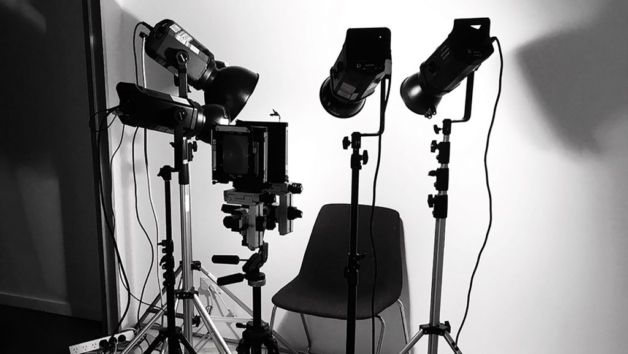 A black and white image of a large format camera and studio lighting set up with a chair in the middle in front of a white backdrop.