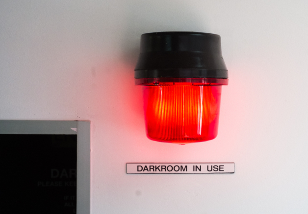 A photograph of a red light fixed to a white wall with a sign underneath reading "Darkroom In Use"
