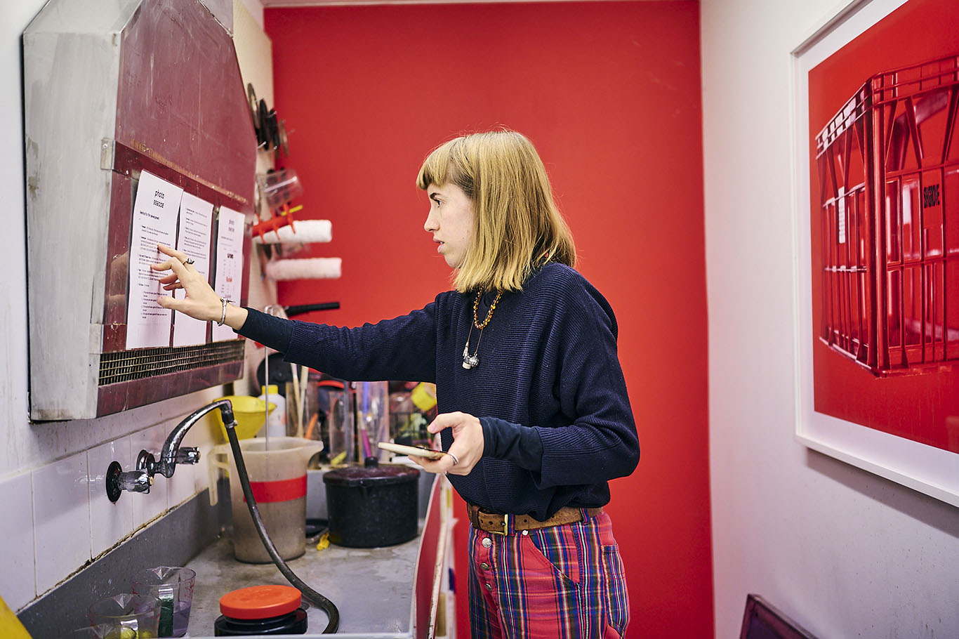 A photograph of a woman in profile, inside a narrow film developing room. She is pointing to some instructions on the wall, with a red painted wall in the background.