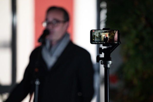 Photograph of an iphone mounted on a tripod to record a man speaking in to a microphone in the background