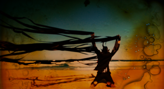 A film still of a man in a black suit and cowboy hat with his arms up and black ribbons flowing in the breeze from his hands behind a sunset background.