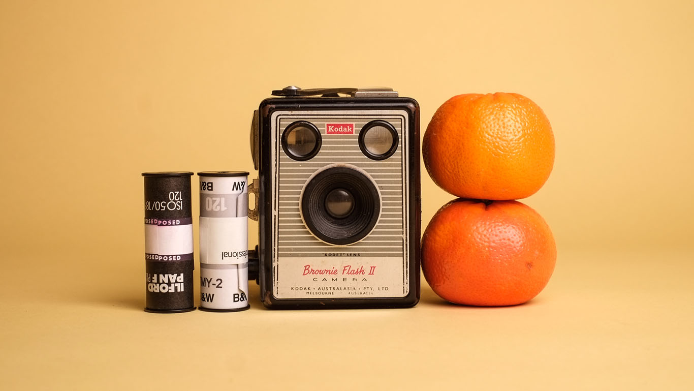A still life arrangement of a medium format camera with two rolls of film on the left and two mandarins on the right, against a yellow paper backdrop.