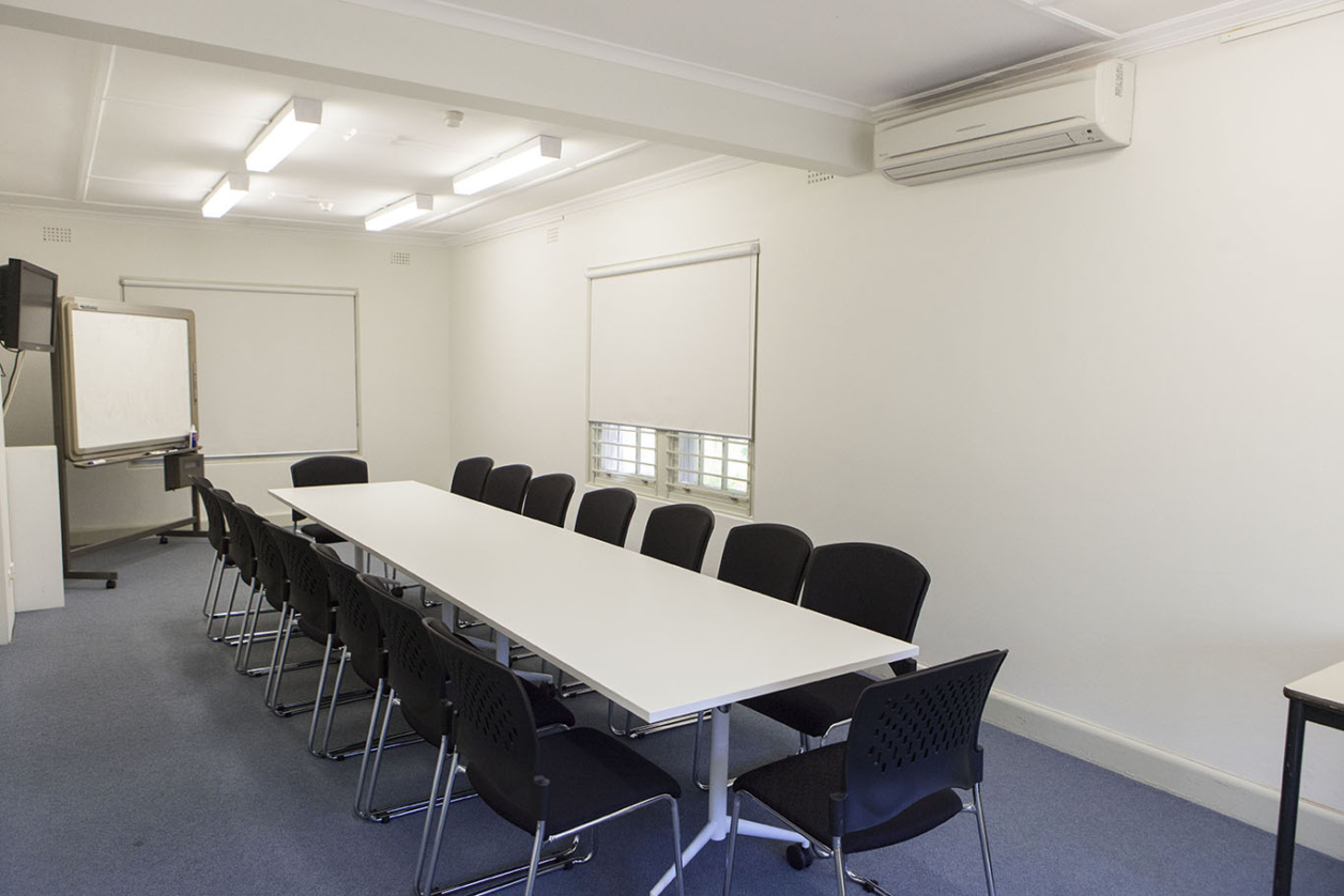 A photograph of a white conference room with a long table and seats around it.