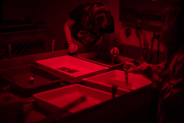 A photograph of two people looking over trays with photographic prints in them in the darkroom.