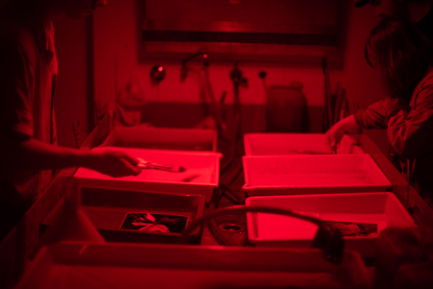 A photograph of hands holding tongs and grabbing onto darkroom trays in a photographic darkroom, all in red light