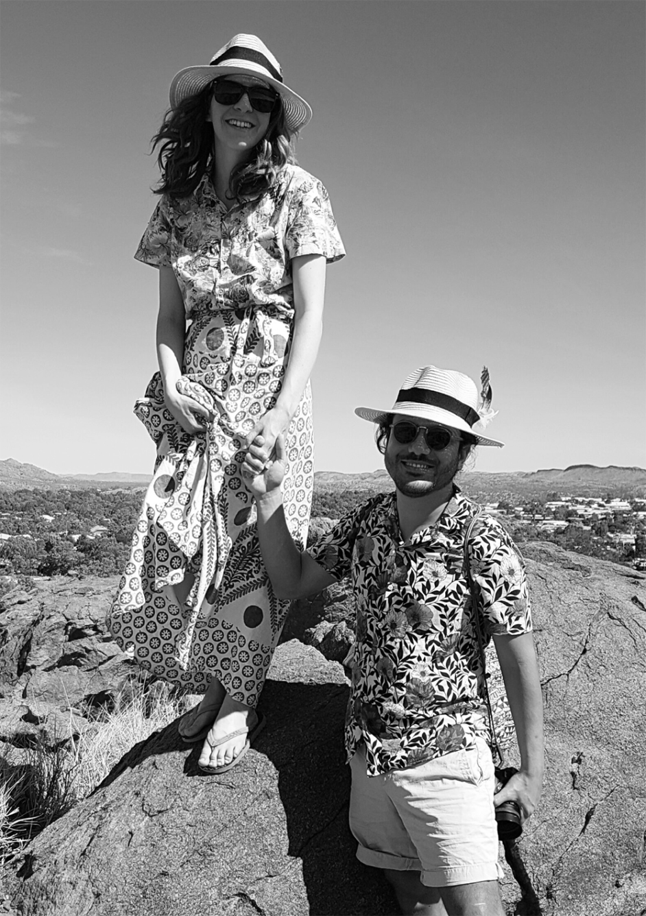 A photograph of a woman standing on a rock holding hands with a man who is below her, smiling to the camera.
