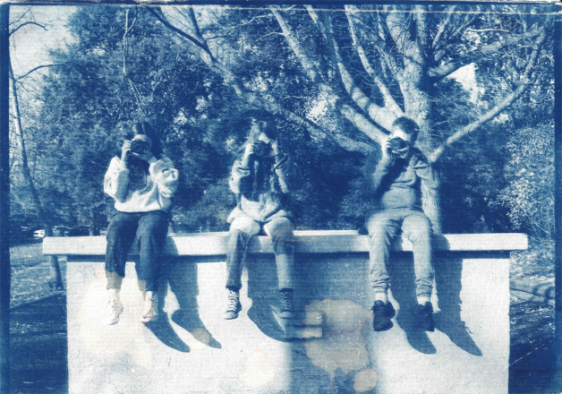A blue and white photograph print of three kids sitting on a ledge with cameras up to their faces facing forwards