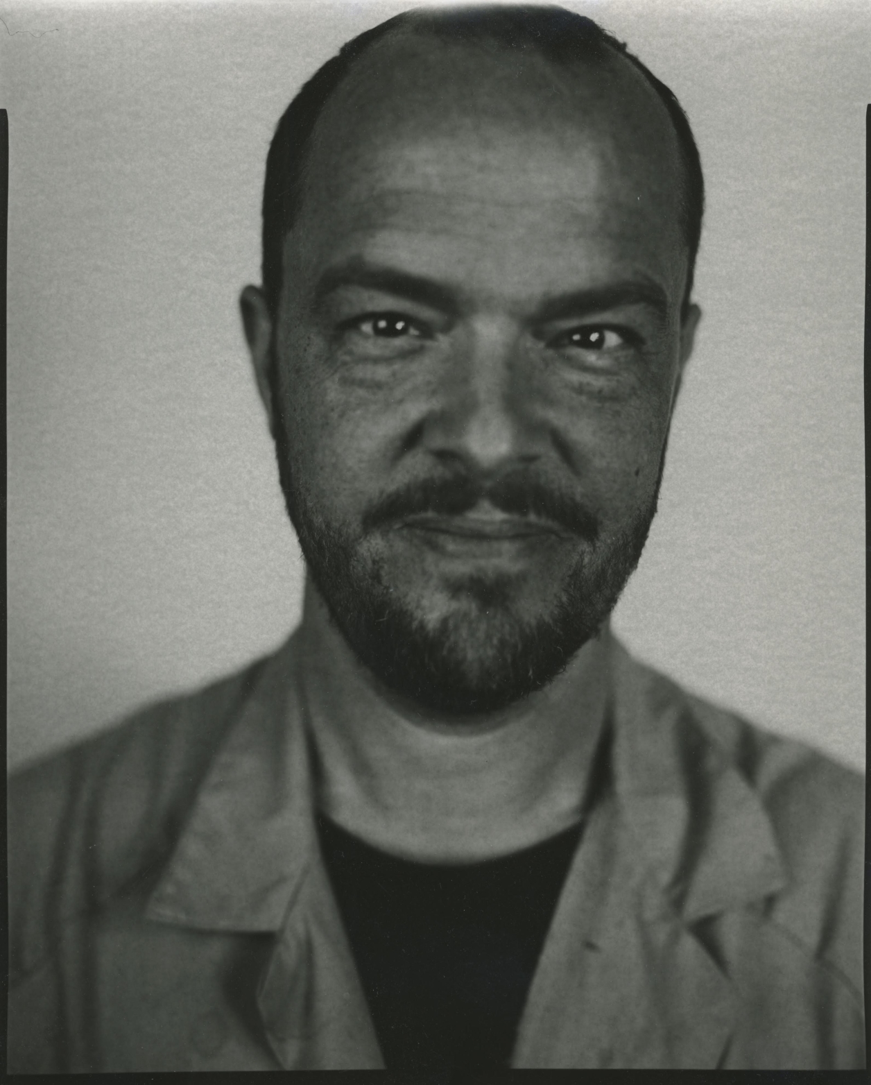 A black and white photograph of a man looking directly at the camera in front of a black background