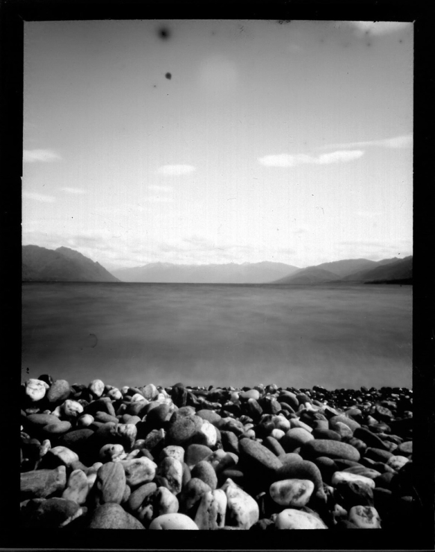 A black and white photograph of a pebbly lake beach with mountains in the background