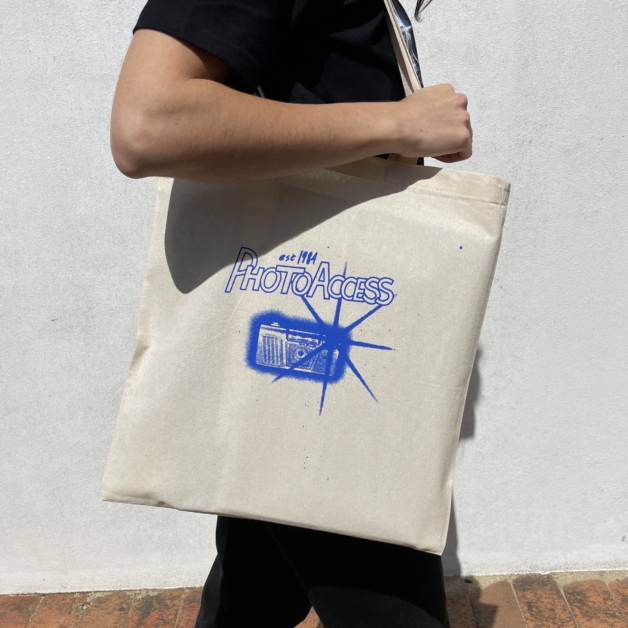 An image of a woman holding a tote bag with a blue camera graphic with a bright flash, and 'PhotoAccess' logo on top