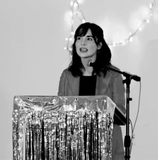 A photograph of a woman with brown hair speaking at a lectern covered in shiny material with lights behind her