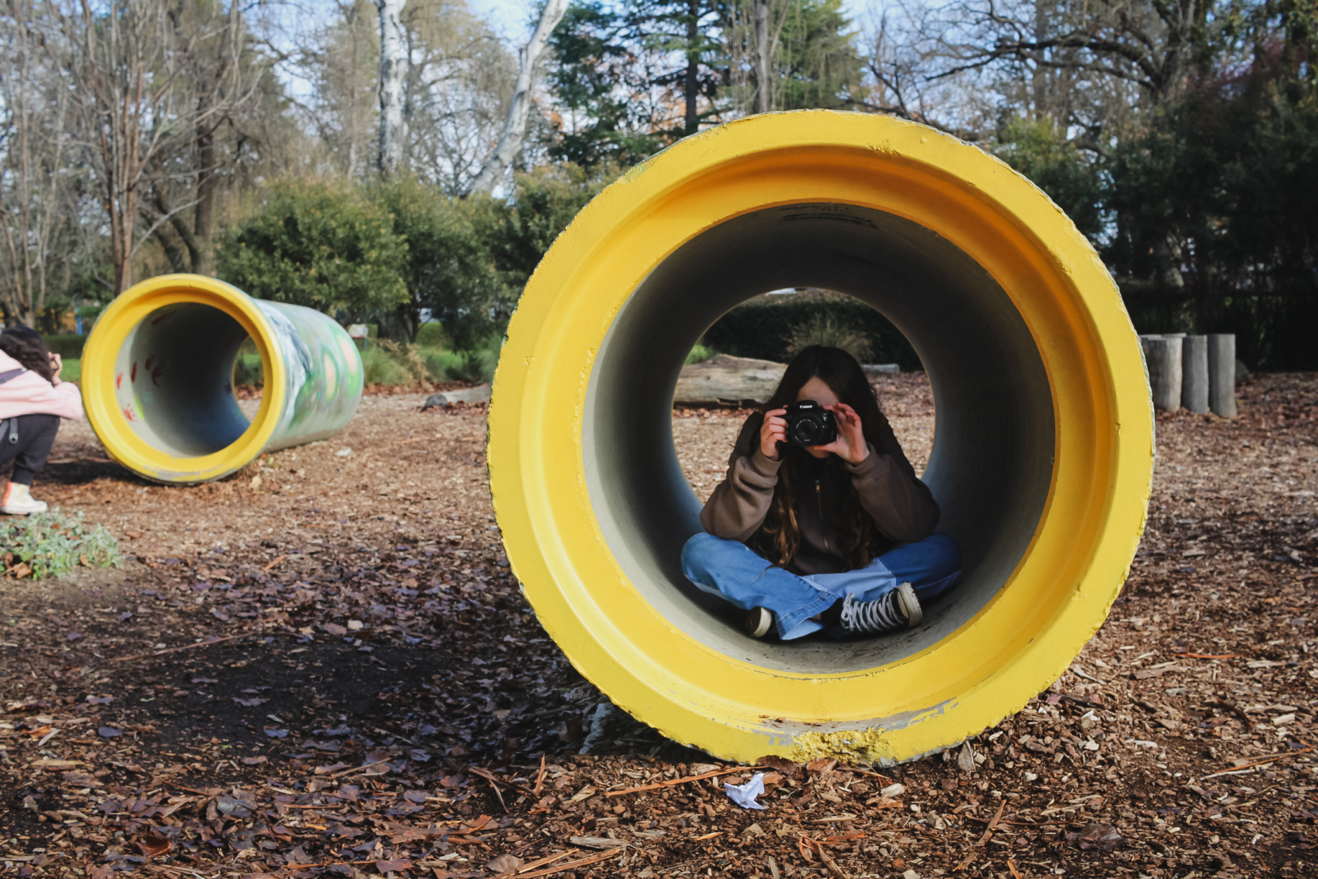 A photograph of a girl sitting in a large yellow pipe holding up a camera taking a photo