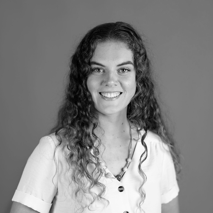 A photograph of a woman with long curly hair smiling at the camera, she wears a white shirt with beaded necklace